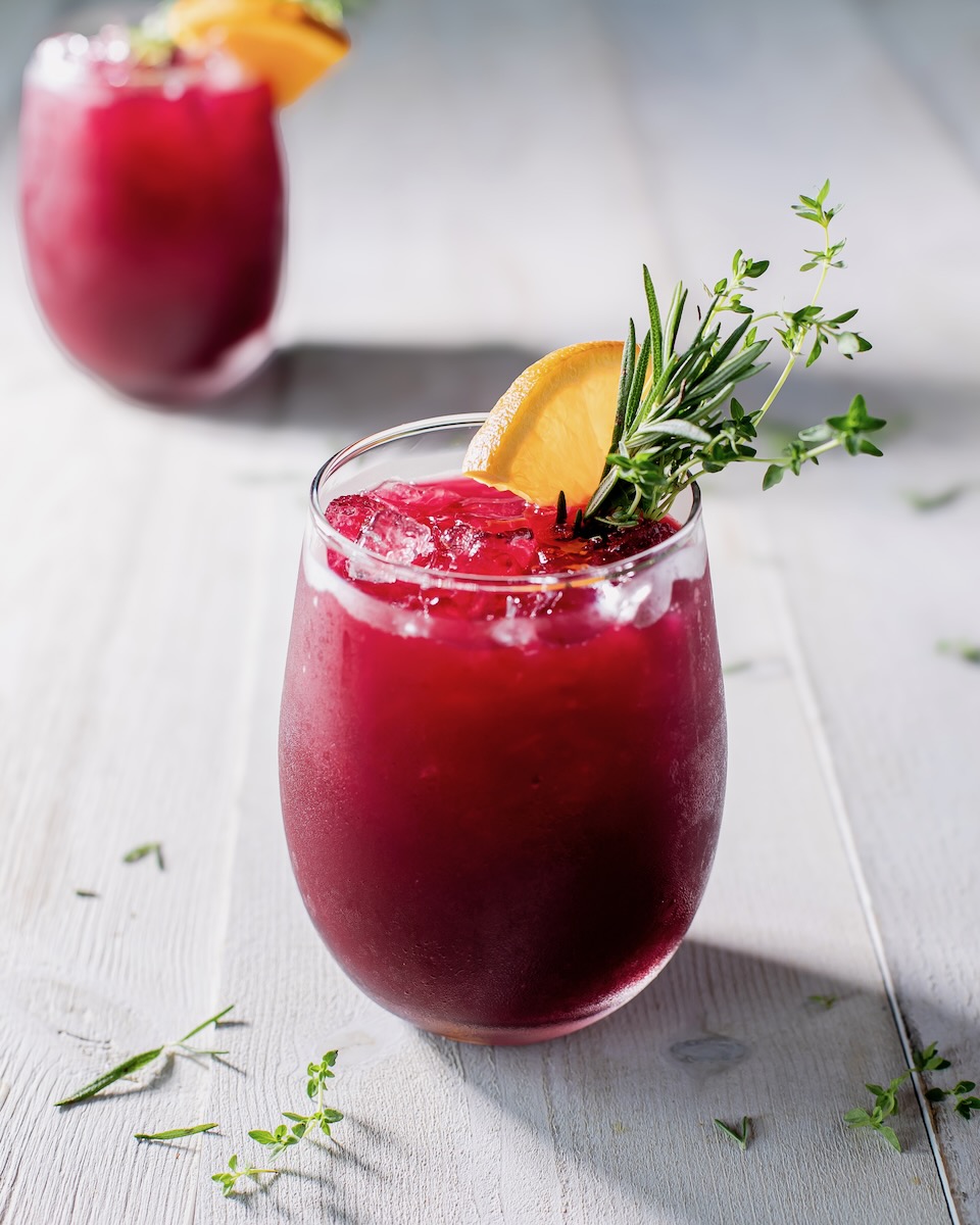 Two cocktail glasses full of beet sangria in a red color sits on a white wood table with orange slices and herb garnishes.