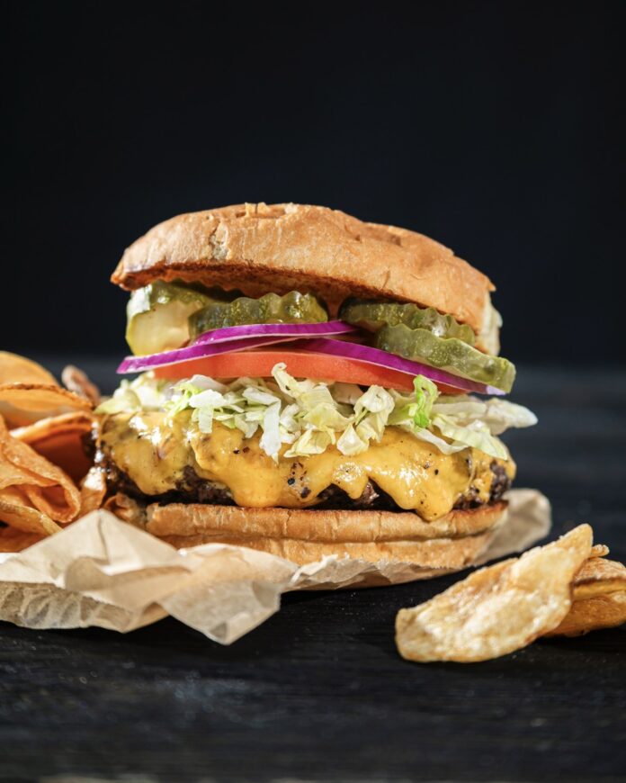 A tall stacked burger with cheese, lettuce, tomato, and pickles with chips around it.