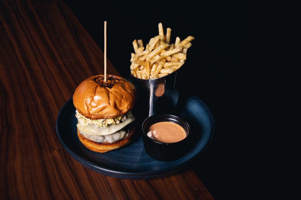 A blue plate with a burger with a pick through it sits next to a silver container of french fries and a pink burger sauce.
