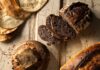 Indulge in a Fresh Loaf of Bread From Pittsburgh Bakeries Various different types of loaves of bread from Pittsburgh bakeries sit on a wood picnic table in light and dark brown colors.