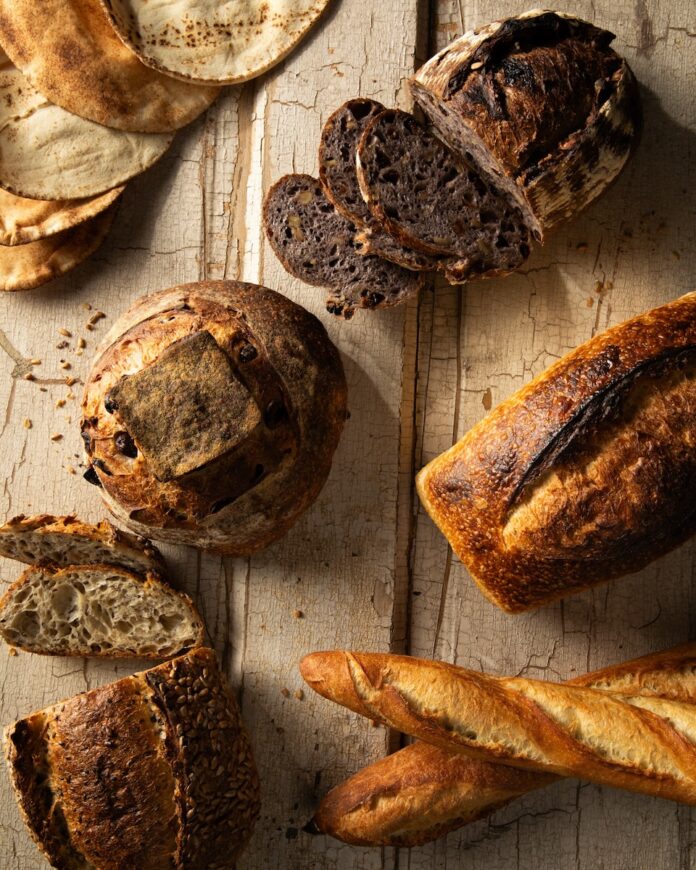 Various different types of loaves of bread from Pittsburgh bakeries sit on a wood picnic table in light and dark brown colors.