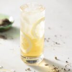 A tall glass with a U.S. Open Court arnold palmer cocktail sits with lemons around the glasses edges and herbs across the table the drink sits on.
