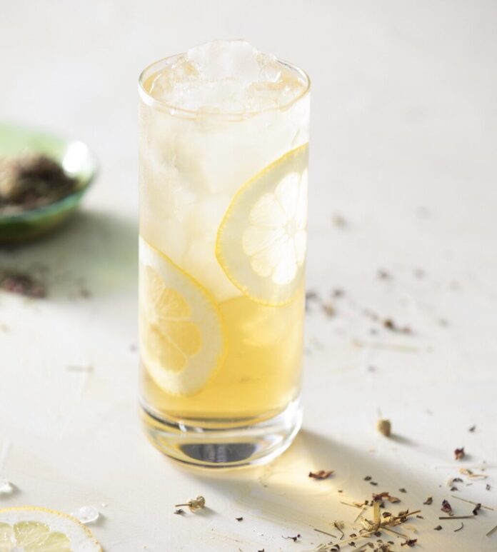The U.S. Open's Arnold Palmer Cocktail A tall glass with a U.S. Open Court arnold palmer cocktail sits with lemons around the glasses edges and herbs across the table the drink sits on.