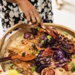 A basket platter holds a variety of charred red cabbage as a woman uses utensils to serve the dish.