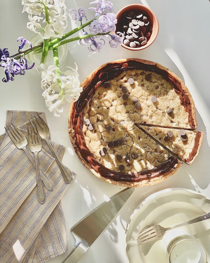 A Chocolate Chip Cake with chocolate ganache on top sits on a white table with white and purple flowers laying near by and a silver serving spatula.