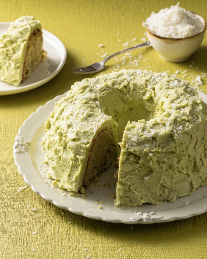 A round chiffon cake with coconut and match buttercream with a slice taken out of it that sits in the background.