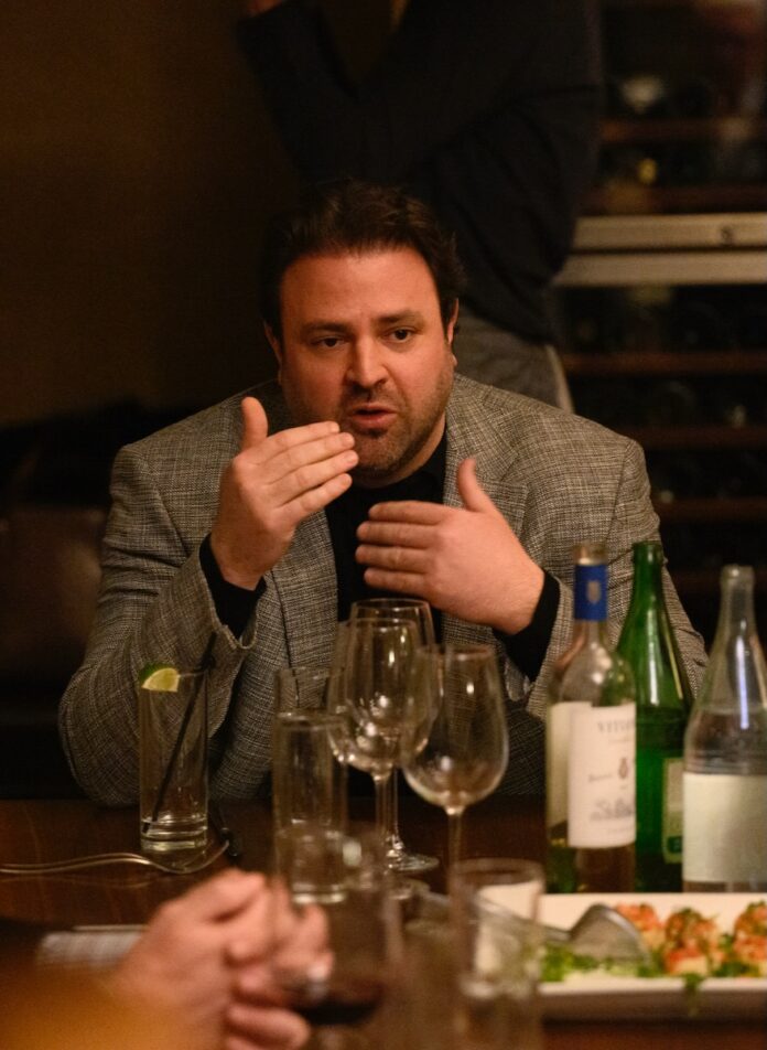 A man in a grey jacket talks with his hands at a dinner table with wine glasses on it.