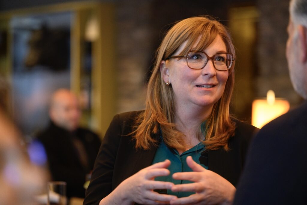 A woman in a black jacket and glasses talks with her hands.