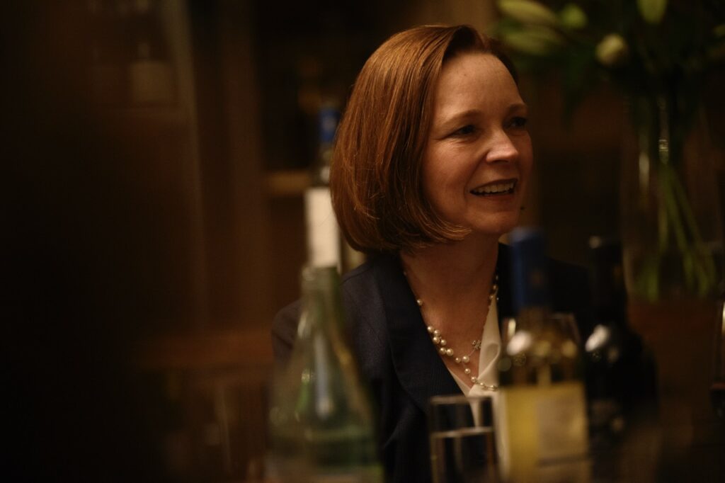 A man with a necklace and black blazer sits at a dining table by wine bottles and glasses.
