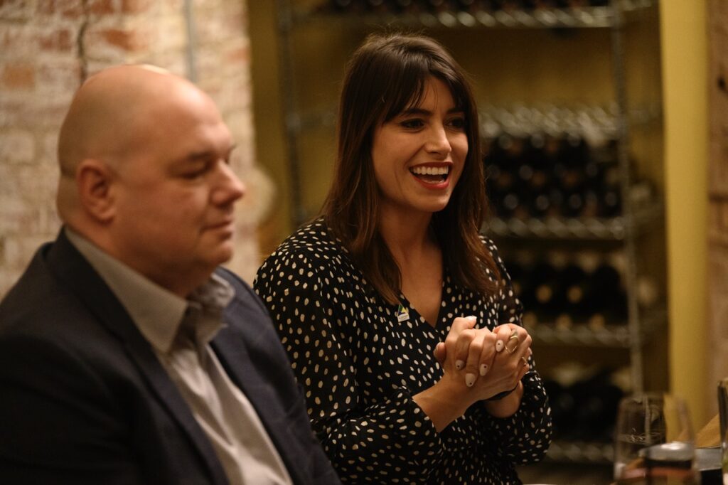 A woman in a polka dot black and white shirt smiles and clasps her hands at a dining table.