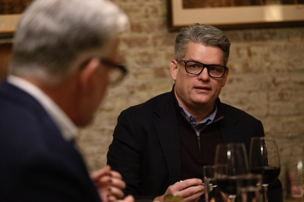 Two men with grey hair and black glasses talk to each other at a dining table.