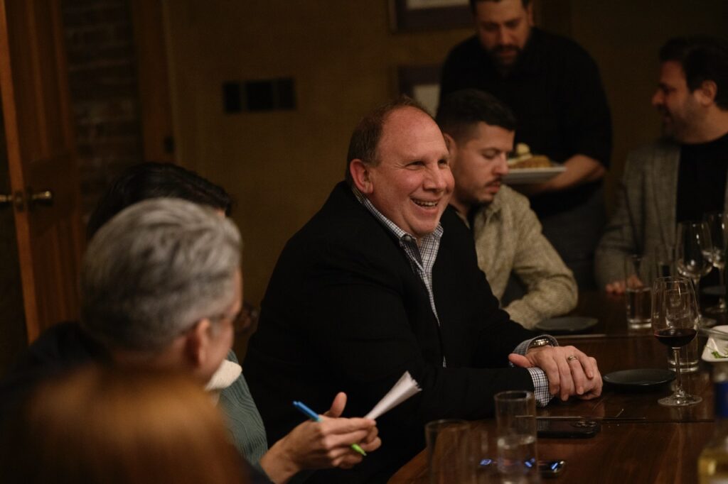 A man in a black jacket smiles at a wood dining table among other Pittsburgh citizens.