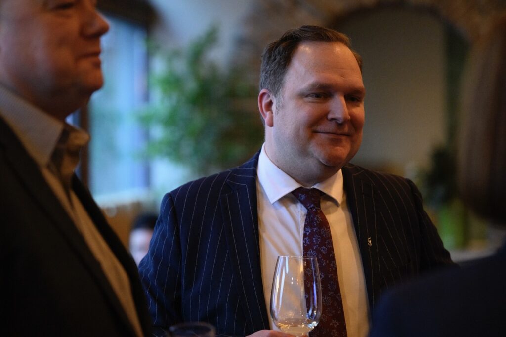 A man in a suit jacket and tie stands with a wine glass in his hand.