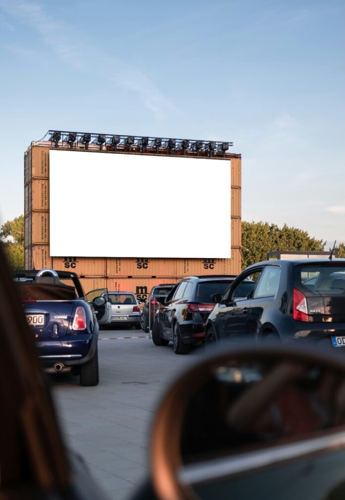 A bunch of cars sit outside of a screen at the drive-in movies.