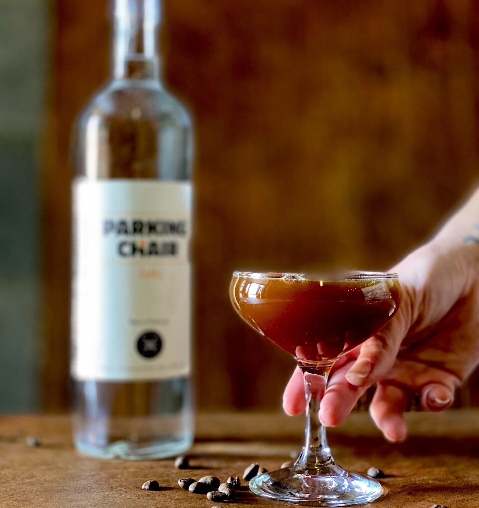 A person places down a coupe glass with an espresso martini in it as a bottle of parking chair vodka sits blurred in the background.