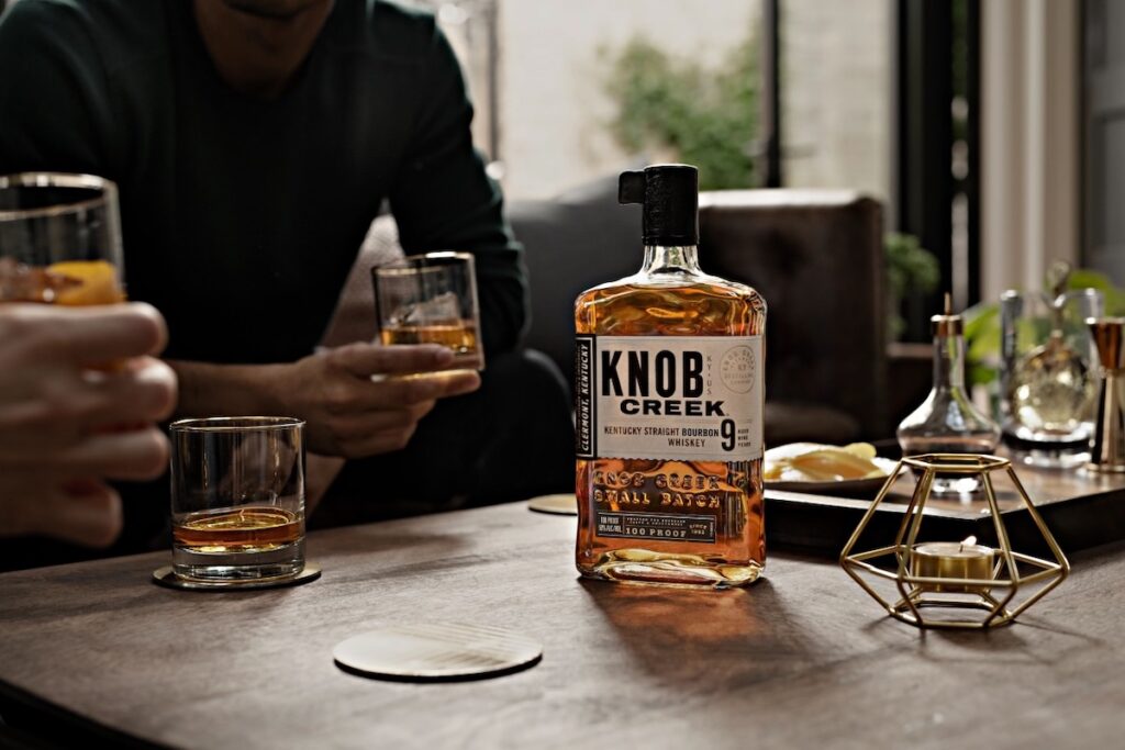A bottle of Knob Creek Whiskey sits on a table as two men enjoy glasses of it.
