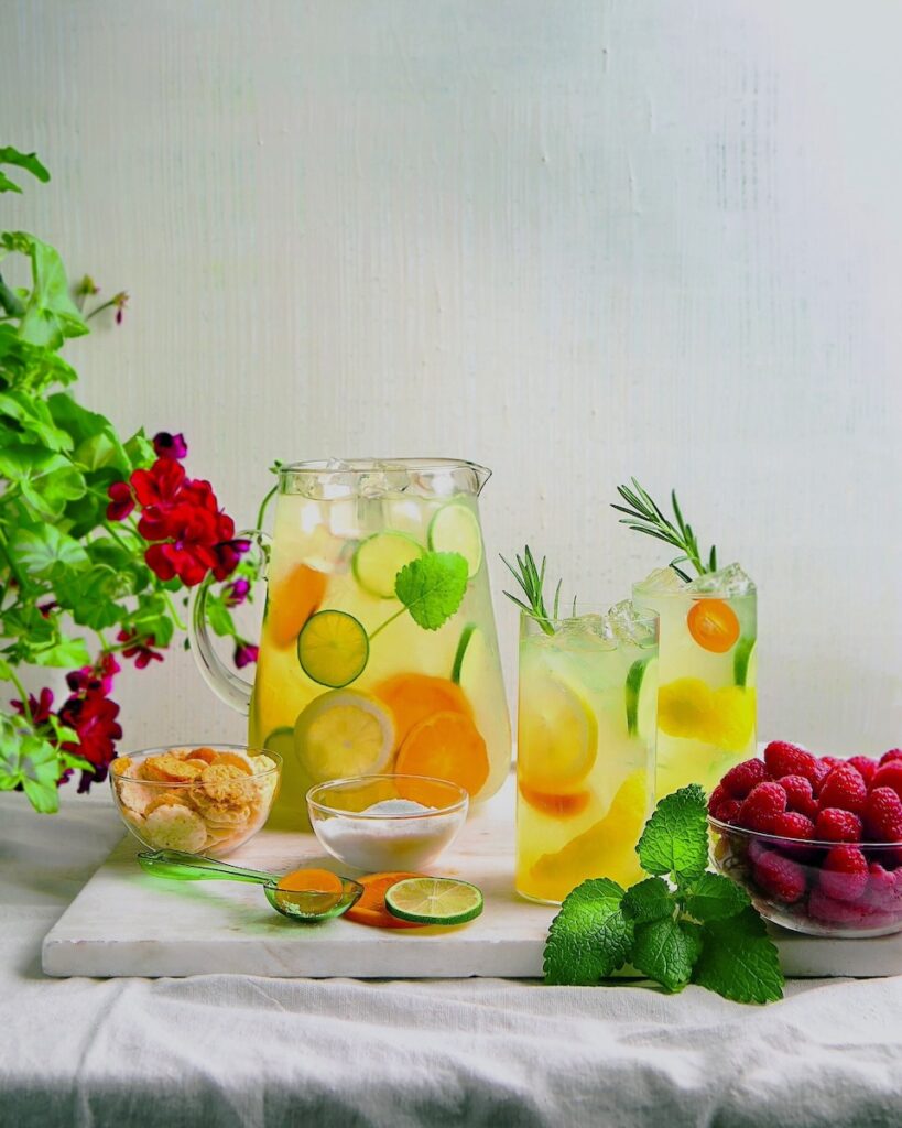 Two glasses of Fresh Summer Lemonade with oranges and limes sit in front of a pitcher of the lemonade as vines of green come in from the sides.