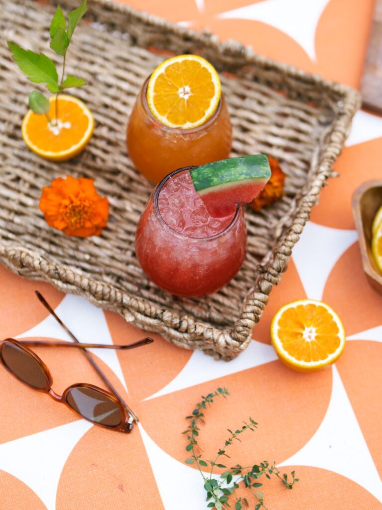 A red watermelon and orange spritz sit on wicker tray on an orange picnic table.