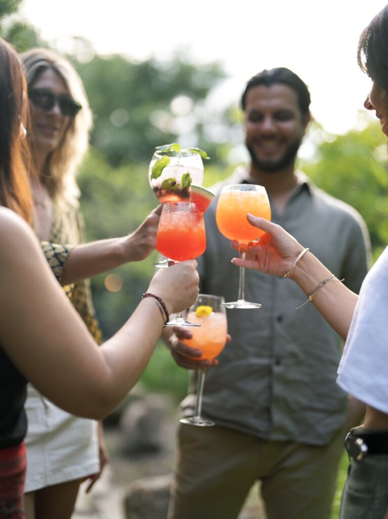 Four people cheer glasses of summer garden-fresh spritzes in orange, red, and white colors.
