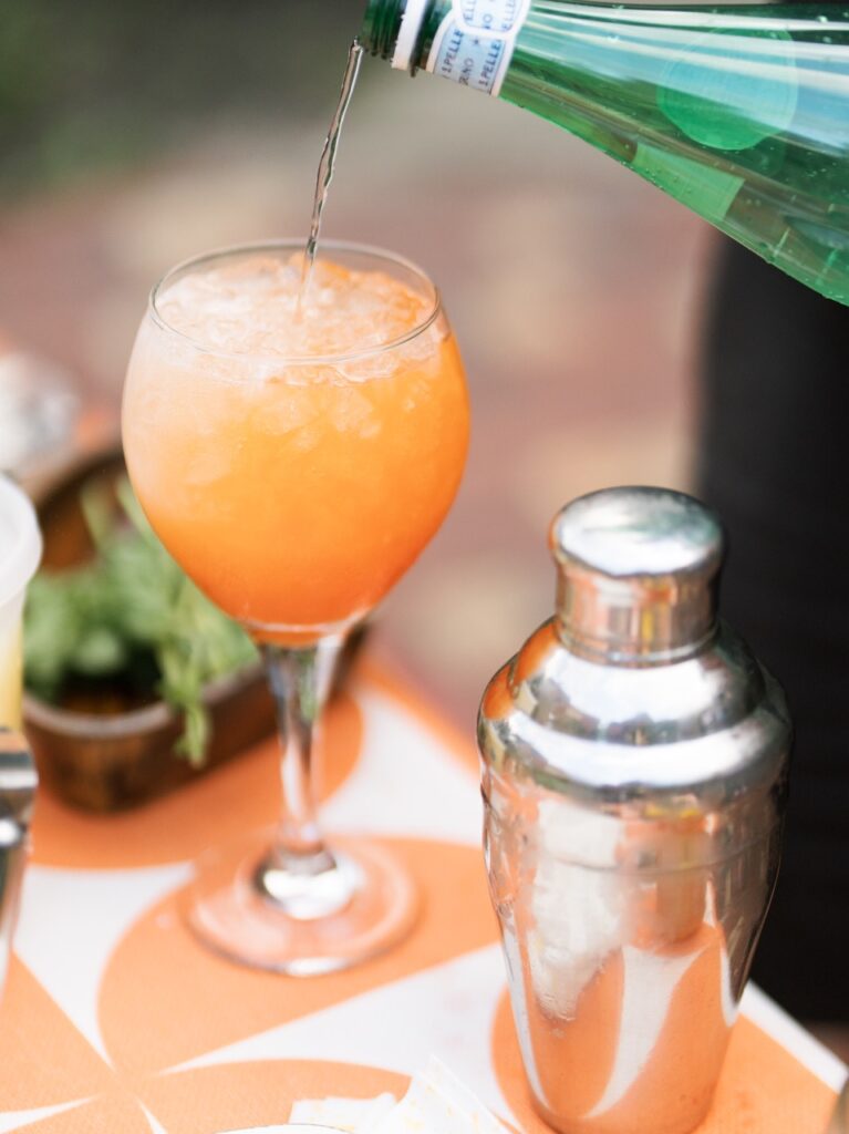Sparkling water is poured into a wine glass to make an orange garden-fresh spritz beside a clear cocktail shaker.