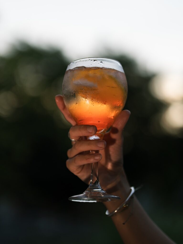 A person holds a glass of summer garden-fresh spritz in the air against a trees background.