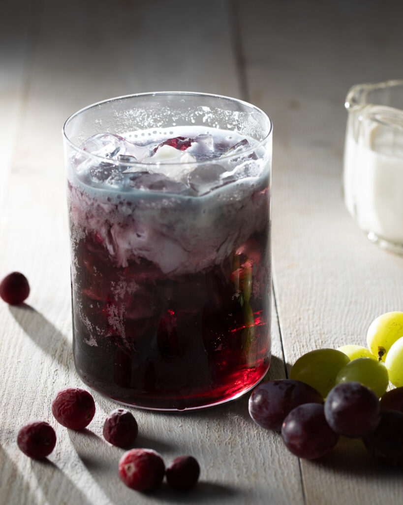 A glass of grape soda with half and half foam on top sits behind a pile of cranberries and grapes as a small container of half and half sits in the background.