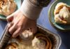 Homemade Cinnamon Rolls Four cinnamon rolls sit in a silver pan as a person reaching in to grab one. Two other cinnamon rolls sit on green plates in the top two corners.