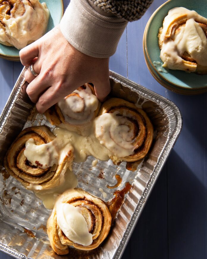 Four cinnamon rolls sit in a silver pan as a person reaching in to grab one. Two other cinnamon rolls sit on green plates in the top two corners.