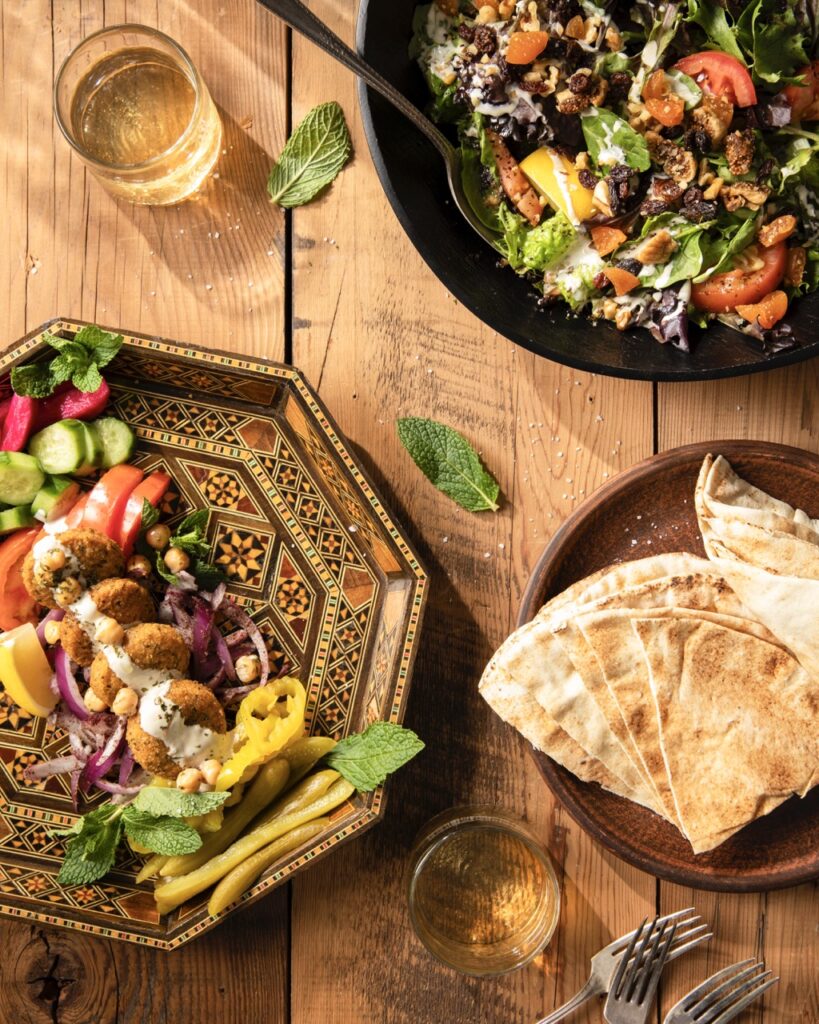 Various plates of international cuisine like veggies in sauce and naan sit on brown plates on a picnic table.