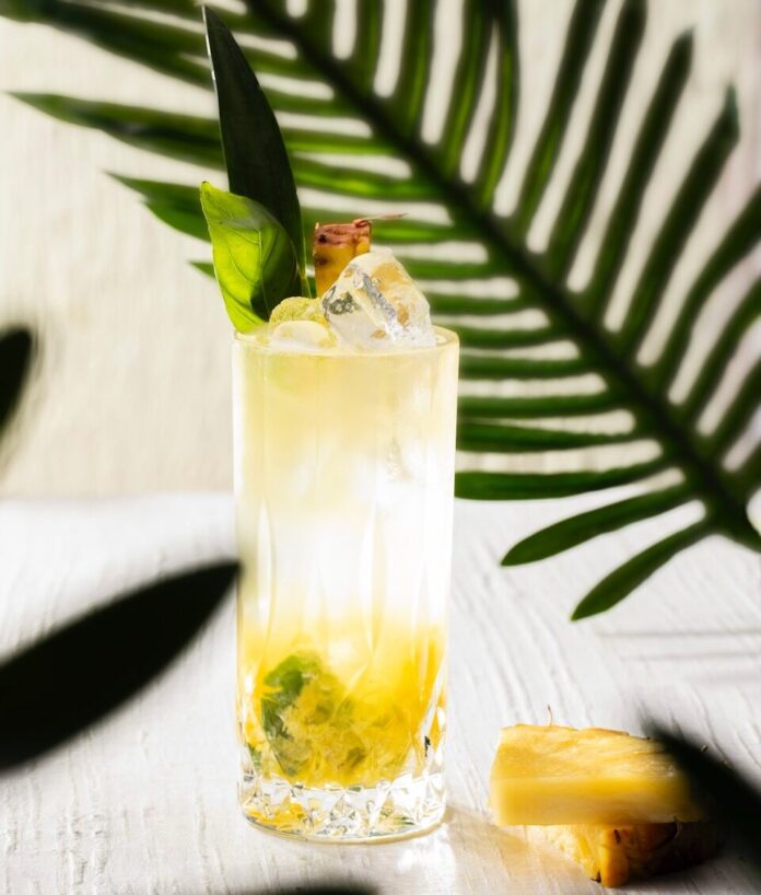 A tall tropical cocktail for Jurassic World: Rebirth with pineapple leaves and mint for garnish, hidden amongst jungle palms.