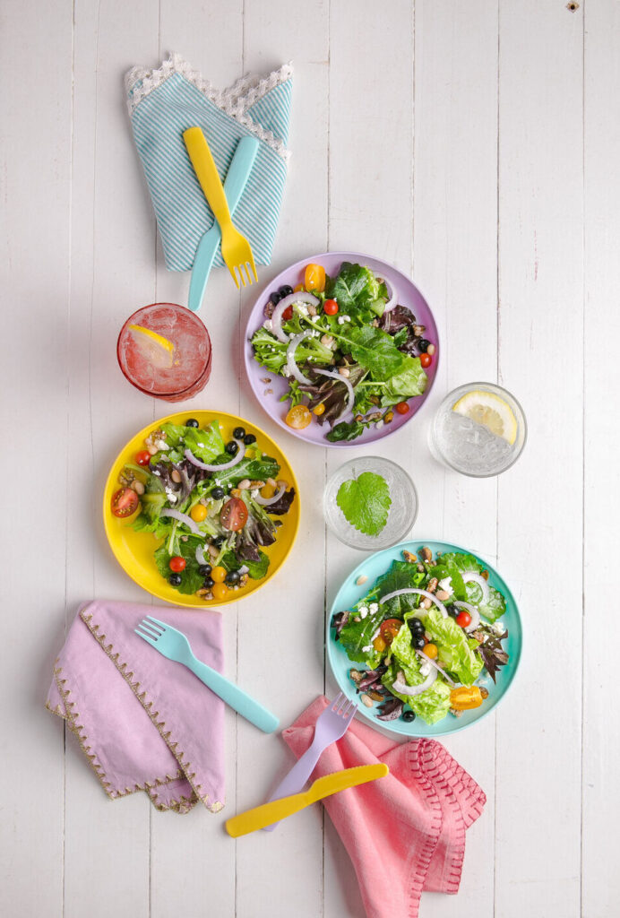 Three colorful bowls of kid-friendly summer salad with pink, purple, and blue napkins on the side.