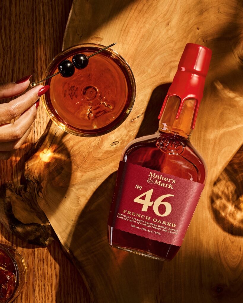 A bottle of Maker's Mark with a red label and seal beside a red cocktail with two black olives.