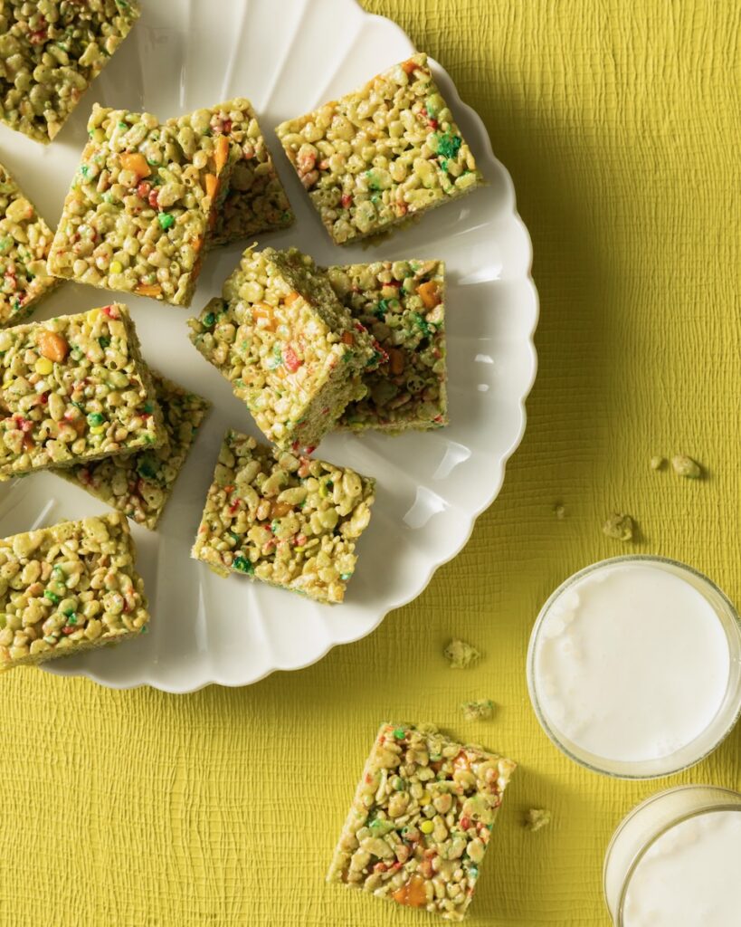 A white plate full of matcha rice krispie treats with mango. It sits beside two glasses of milk on a green background.
