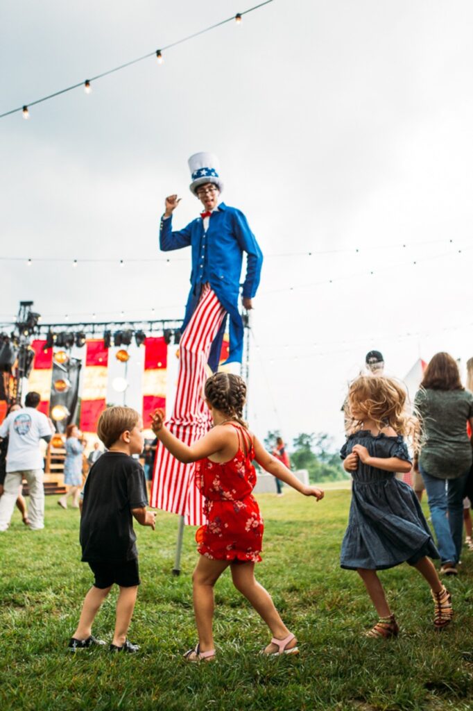 A man with long pinstriped pants and a blue jacket stands on stilts in front of children at a Fourth of July event in Pittsburgh.