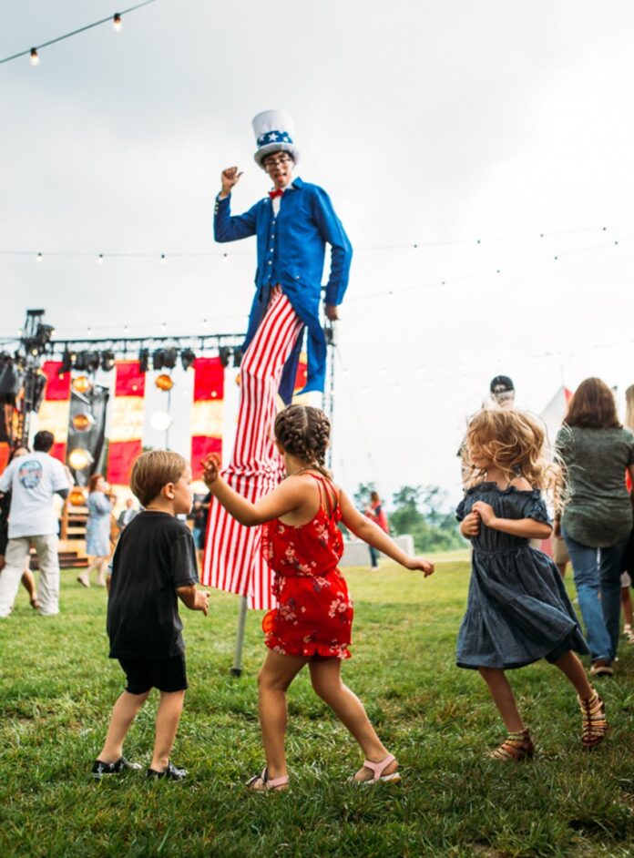 A man with long pinstriped pants and a blue jacket stands on stilts in front of children at a Fourth of July event in Pittsburgh.