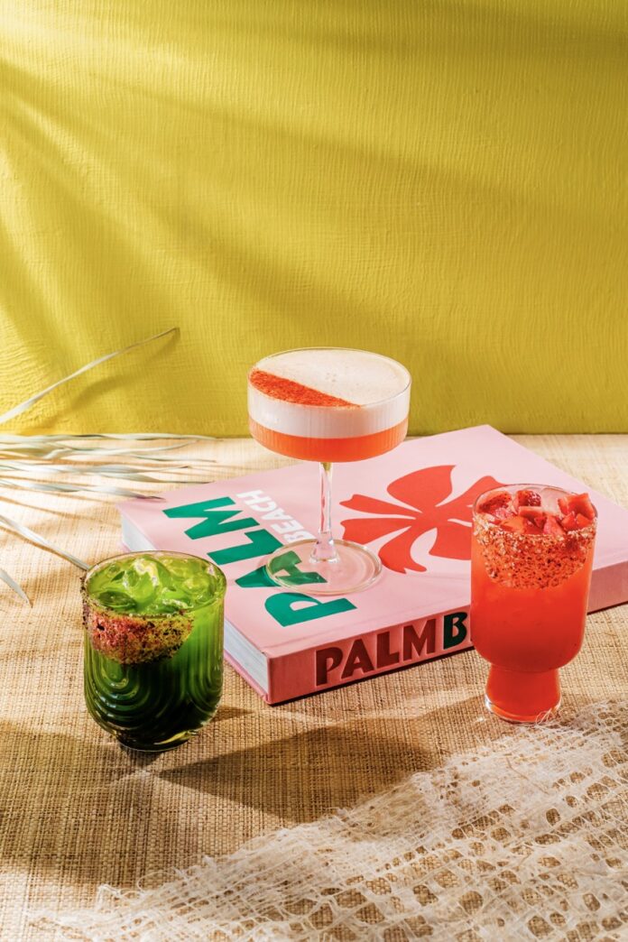 Three cocktails from Palm Palm sit on a brown table with one propped up on a pink Palm book.