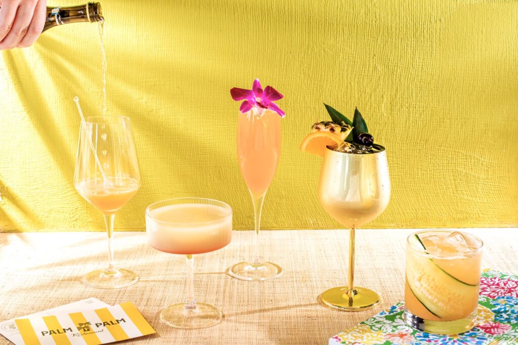 A series of Palm Palm cocktails in various glasses sit in a line across a brown take as a person pours champagne into the first glass.