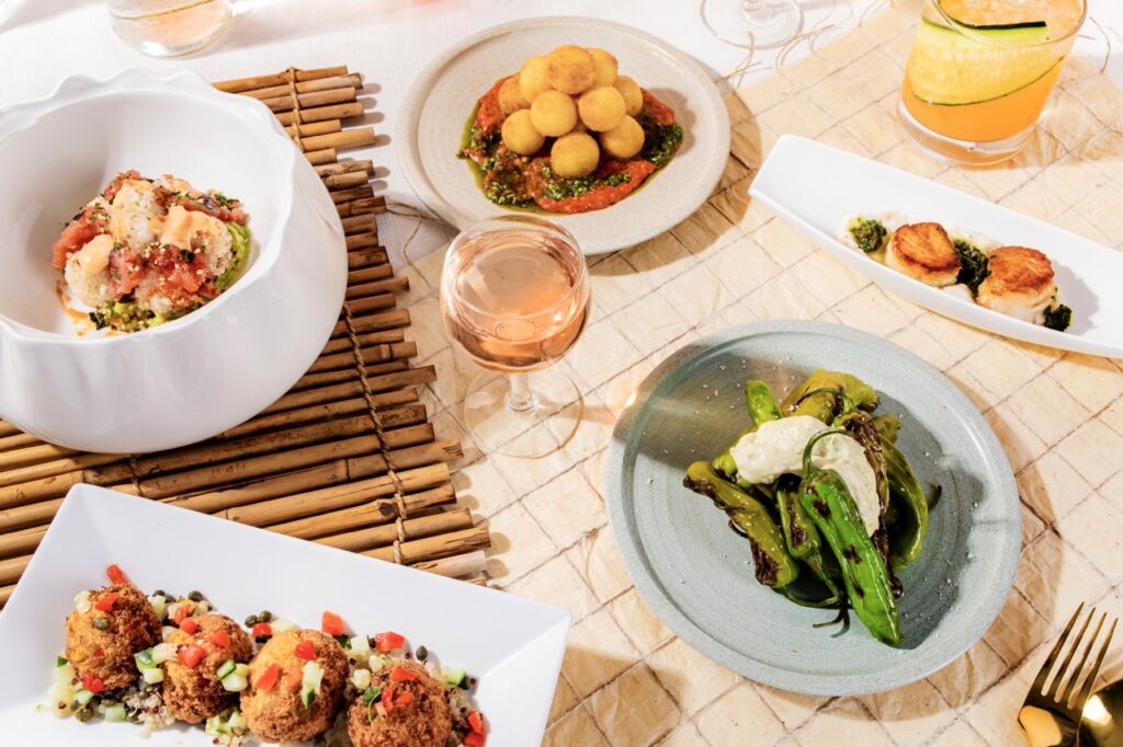 Four white plates and one round blue plate hold appetizers from Palm Palm in Pittsburgh with a glass of wine sitting in the middle of them.
