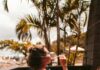 Refreshing Summer Wines for Sipping on the Porch A woman sits on a porch out by the beach sipping a glass of summer wine with palm trees in the background.