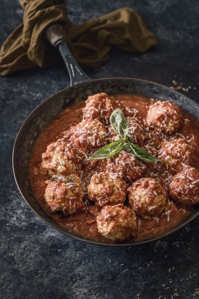 A bunch of ricotta vegetarian meatballs sit in a cast iron pan, soaked in re sauce with basil garnish on top.