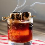 A short glass holds a S'mores Old Fashioned Mocktail featuring a skewer of toasted marshmallows on top and a rim of brown sugar.