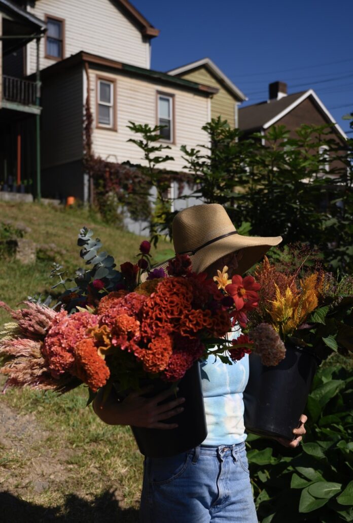 Sol Patch Garden is Braddock's Home for Flowers A woman in a sun hat carries bright red flowers outside a house.
