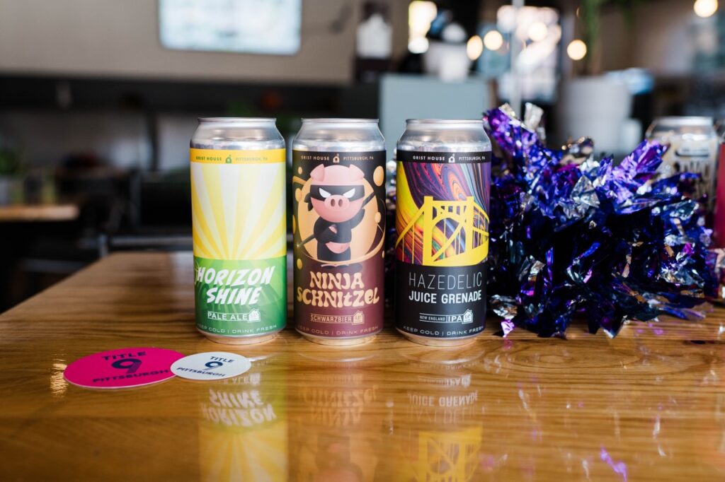 Three cans of beer sit on a wood table beside a pompom at a Pittsburgh sports bar.