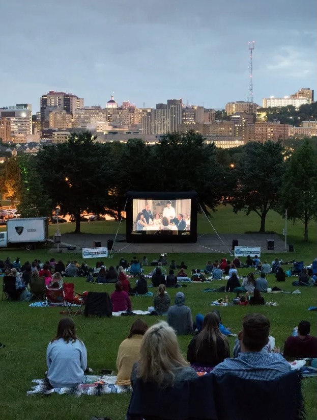 Groups of people on blankets and in chairs sit in a Pittsburgh park watching a free summer movie series on an inflatable screen in front of the city skyline.