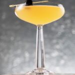 A yellow Superman's Kryptonite cocktail sits in a coupe glass with a toothpick with a black olive on it balancing on the edge. The glass sits on a stone block in front of a silver background.