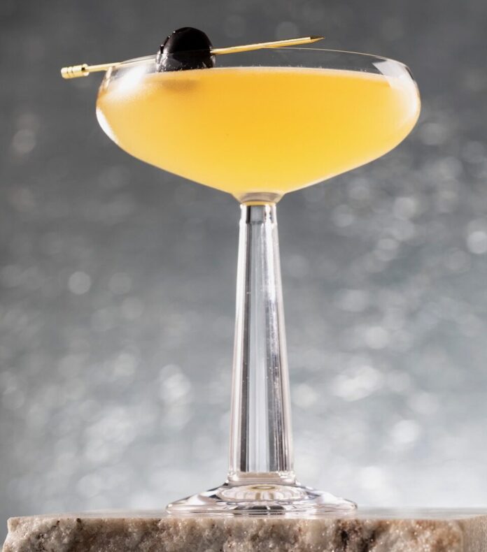 A yellow Superman's Kryptonite cocktail sits in a coupe glass with a toothpick with a black olive on it balancing on the edge. The glass sits on a stone block in front of a silver background.