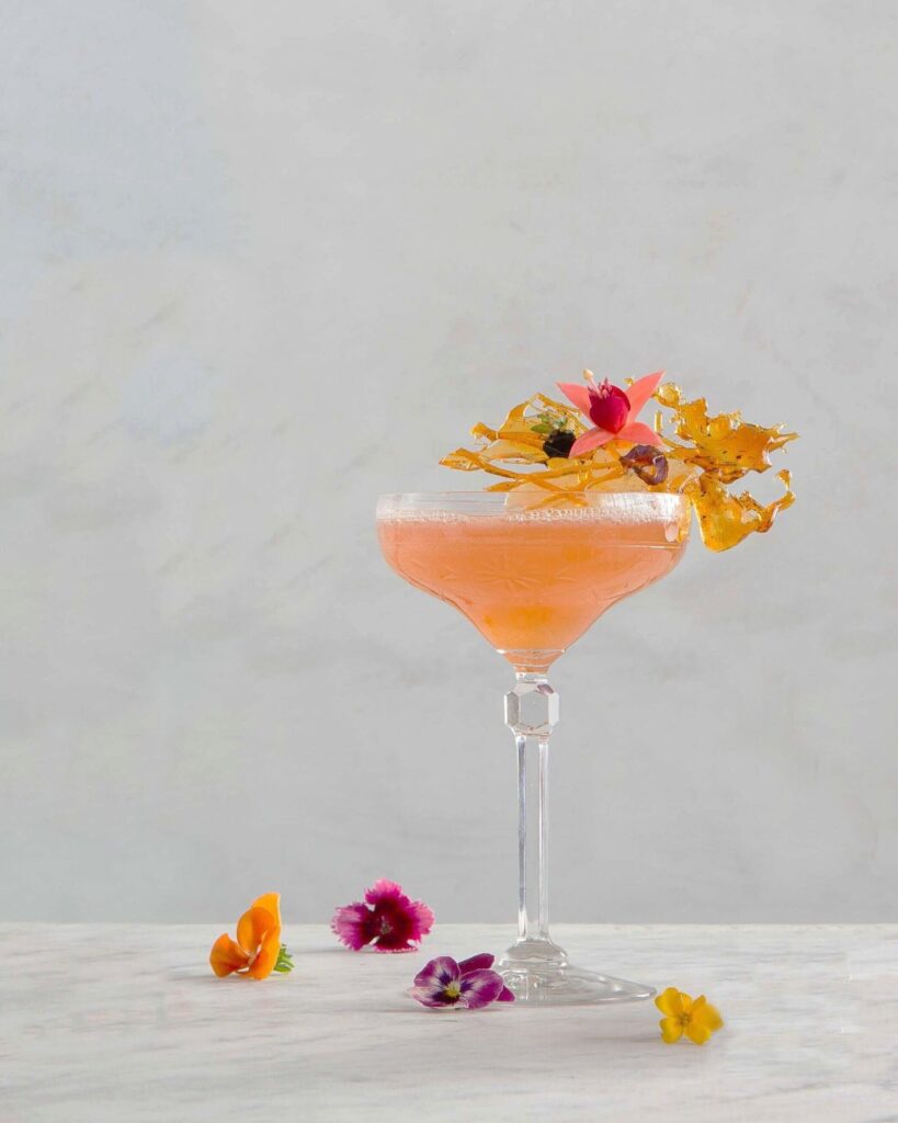 A tarragon gin and yellow chartreuse cocktail sits on a white marble table with edible flowers garnishing the glass and around the table.