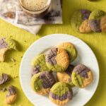 A white plate holds a stack of tricolor matcha sugar cookies in orange, purple, and green. The plate sits on a green background with a coffee above the plate and broken sugar cookies around it.