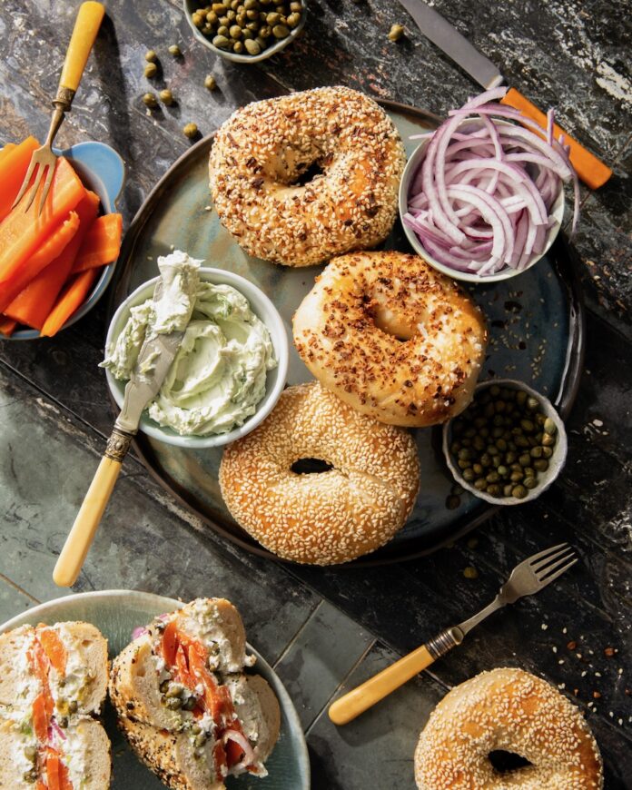 A plate of bagels with various spreads and vegetables around hem sit on a dark wood table.