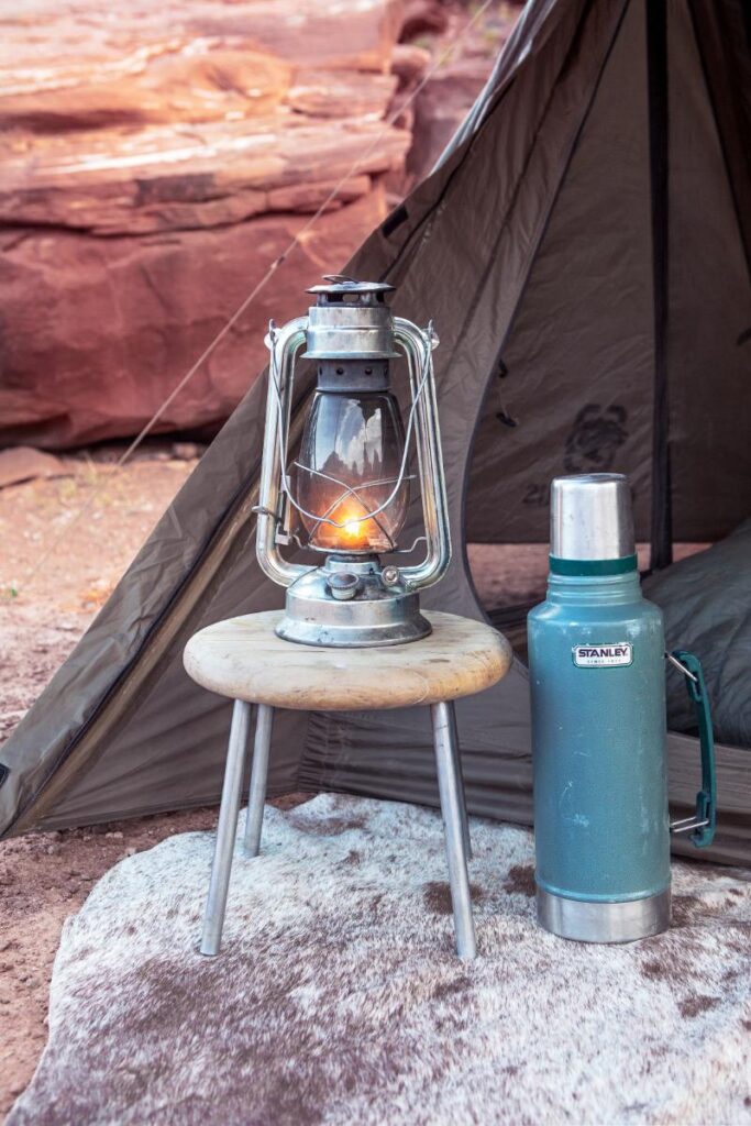 A lit lantern sits on a small wooden stool next to a green thermos and a brown camping tent.